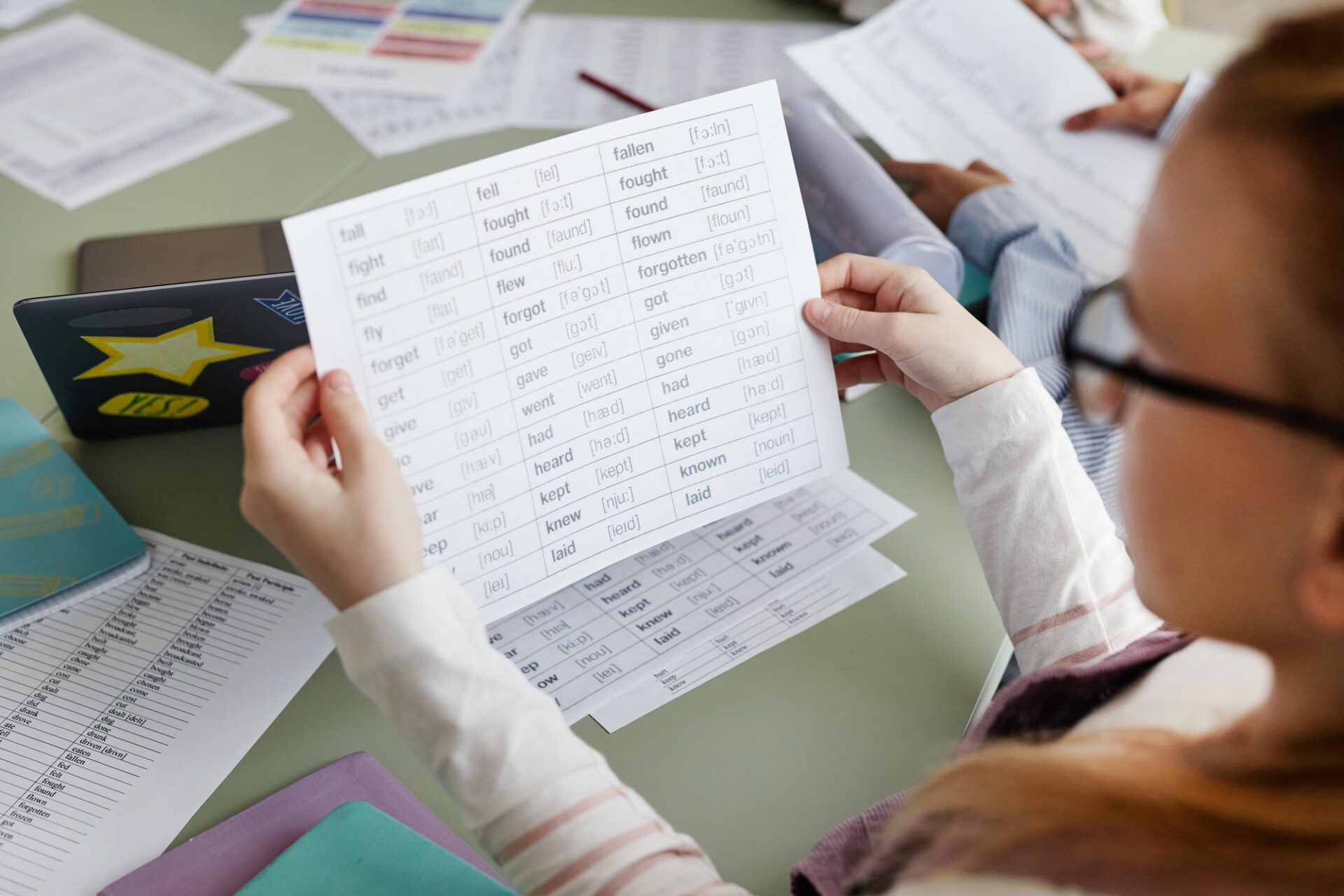 Vue à angle élevé de la fille portant des lunettes assises à table dans la salle de classe Holding Paper Liste de verbes irréguliers